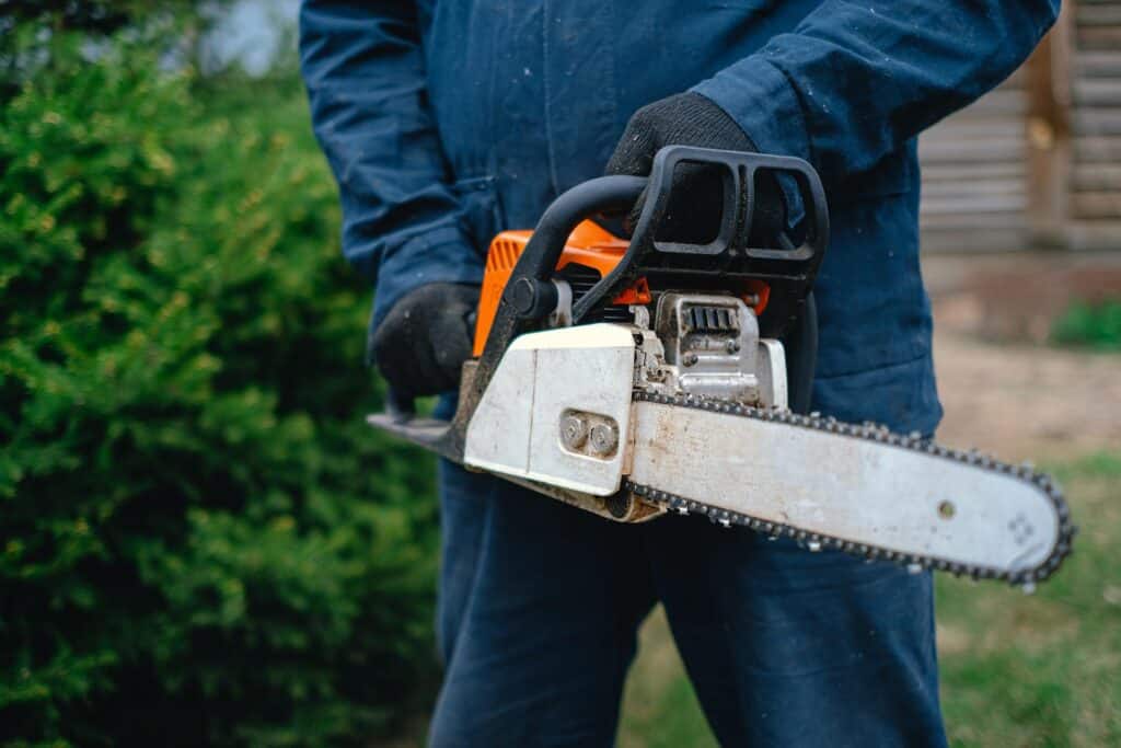 A worker in protective gear holds a chainsaw, preparing for tree trimming. The person is wearing dark gloves and a navy-blue work suit, standing outdoors near greenery.